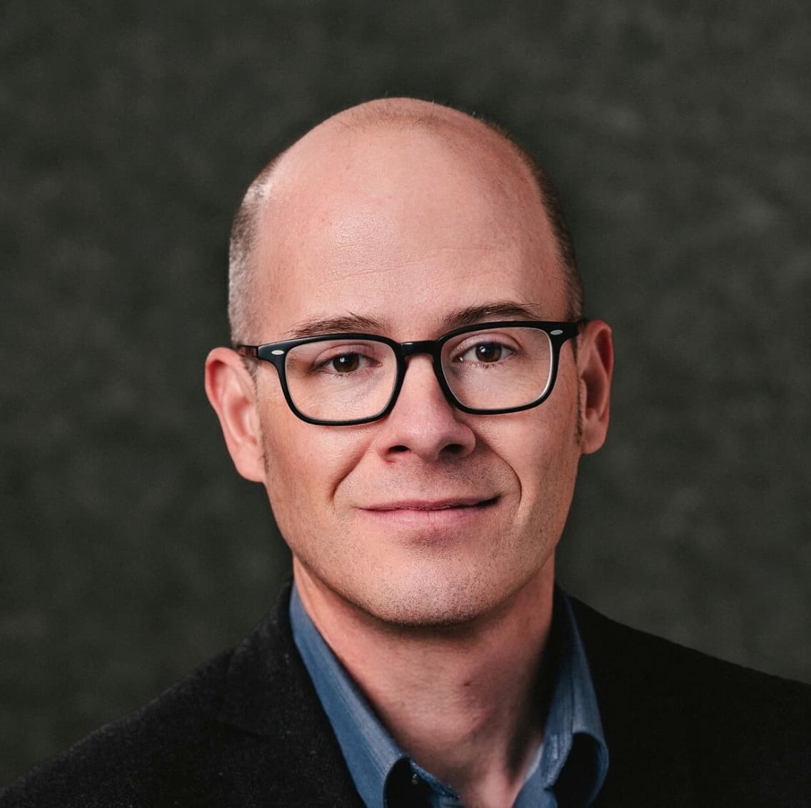 Headshot of a middle-aged man with a bald head wearing black rectangular glasses, a dark blazer, and a blue collared shirt against a dark blurred background.